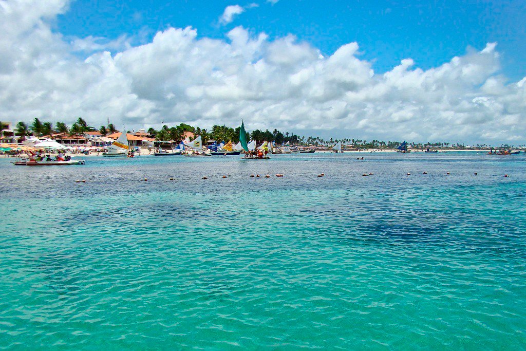 Praia de Porto de Galinhas, água cristalina e no fundo barcos a vela e a praia.