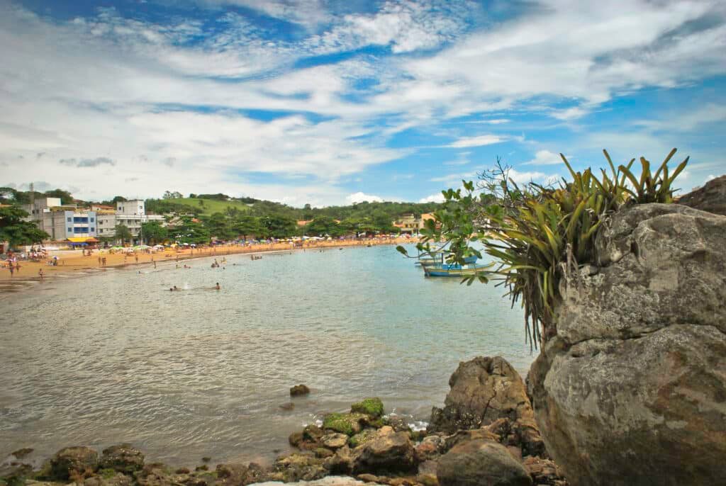 Praia de Iriri, no Espírito Santo. Uma pedra no canto direito com plantas em cima, o mar atrás e no fundo a areia com pessoas no mar.