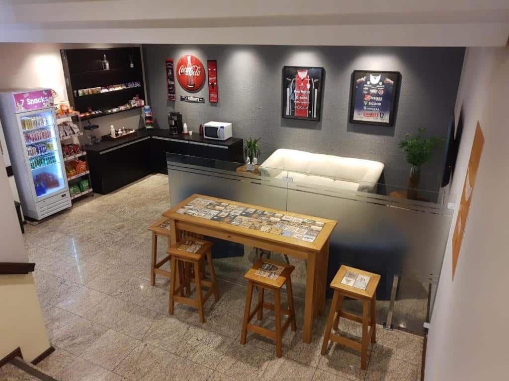 Área de convivência do 7HOLZ mostrando um sofá branco à direita, com uma mesa pequena de quatro lugares e no canto esquerdo com uma bancada de madeira preta, equipada com microondas e cafeteira elétrica.