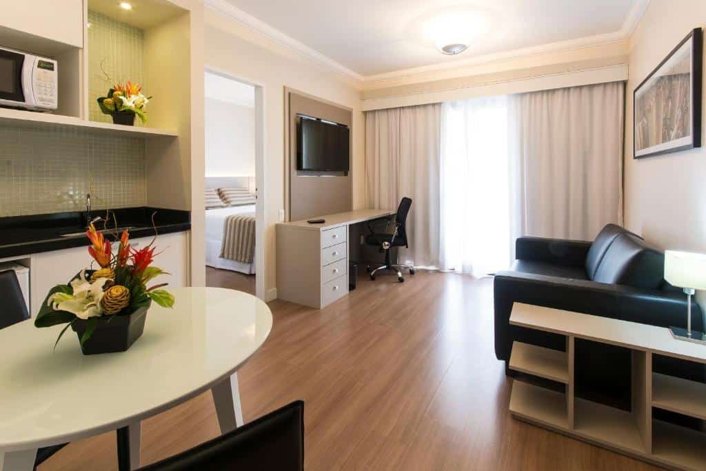 Visão da suíte do hotel. Há uma cozinha equipada com gabinete, uma pia, micro-ondas e armário. Há uma mesa branca para refeições com duas cadeiras pretas e um vaso preto com flores no centro. Na sala, há um sofá preto, uma mini estante do lado com um abajur e um quadro em cima. Na frente, uma TV suspensa na parede e uma escrivaninha branca com gavetas e uma cadeira giratória preta. Do lado do sofá e da escrivaninha, está a janela, com uma cortina de cor clara.