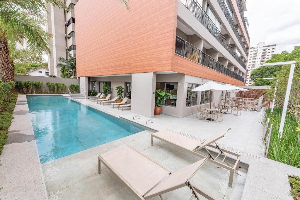 Área externa de lazer do prédio do Charlie Vitrali em Moema. Temos a visão da piscina com várias espreguiçadeiras em tom de madeira clara em volta, e algumas mesas com guarda-sol e cadeiras. Também há um jardim em volta de todo o terreno, com plantas e árvores.