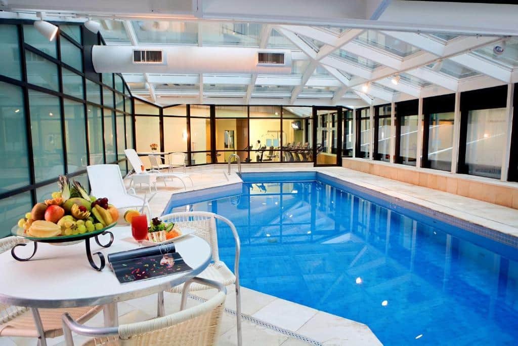 Área da piscina interna coberta do hotel Bourbon, é separada por um vidro com a academia do hotel. Há uma mesa com frutas e várias espreguiçadeiras ao redor da piscina. Foto para ilustrar o post de hotéis em Moema.