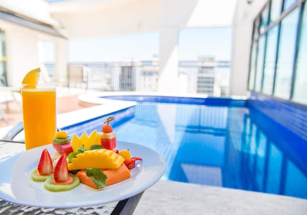 Piscina interna do hotel Estanplaza (sem cobertura). Há espreguiçadeiras em volta da piscina. No primeiro plano da foto, um prato com frutas e um suco de laranja está em foco.