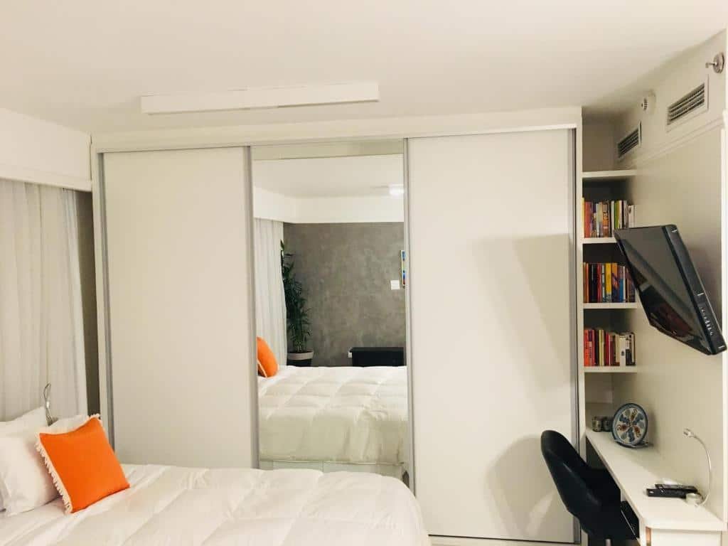 Visão do quarto do hotel. A cama é de casal, com travesseiros brancos e um laranja, o lençol também é branco. As cortinas atrás da cama são brancas. O guarda-roupa é espelhado e é tamanho de casal, também branco. Possui uma estreita estante com alguns livros do lado direito. De frente para a cama, há uma pequena escrivaninha branca, com uma cadeira preta, e uma TV está fixa na parede. Foto para representar o post sobre hotéis em Moema.