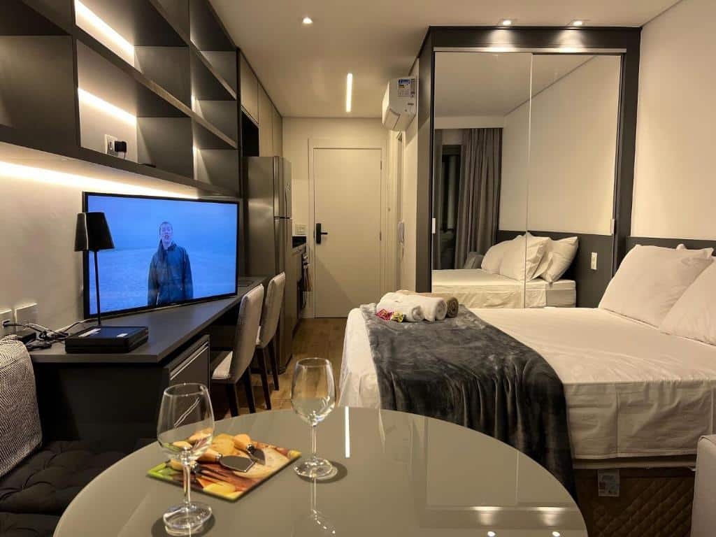 Vista do apart-hotel do Studios Sampa. Vemos uma mesa cinza, com duas taças e utensílios de cozinha em cima sobre um prato colorido. Há um pequeno sofá preto encostado na parede que acompanha a mesa. Do lado do sofá, há uma escrivaninha preta com uma TV e abajur em cima, comporta duas gavetas debaixo. Há duas cadeiras cinzas almofadadas que acompanham a escrivaninha. De frente, tem uma cama box de casal com lençóis brancos, travesseiros brancos, uma manta cinza por cima e algumas toalhas de banho dispostas pelo serviço de quarto. Do lado da cama, há um guarda-roupa espelhado. Perto da porta de entrada, tem uma cozinha equipada e de frente para ela, um banheiro privativo. O interfone está do lado da porta do banheiro, fixo na parede.