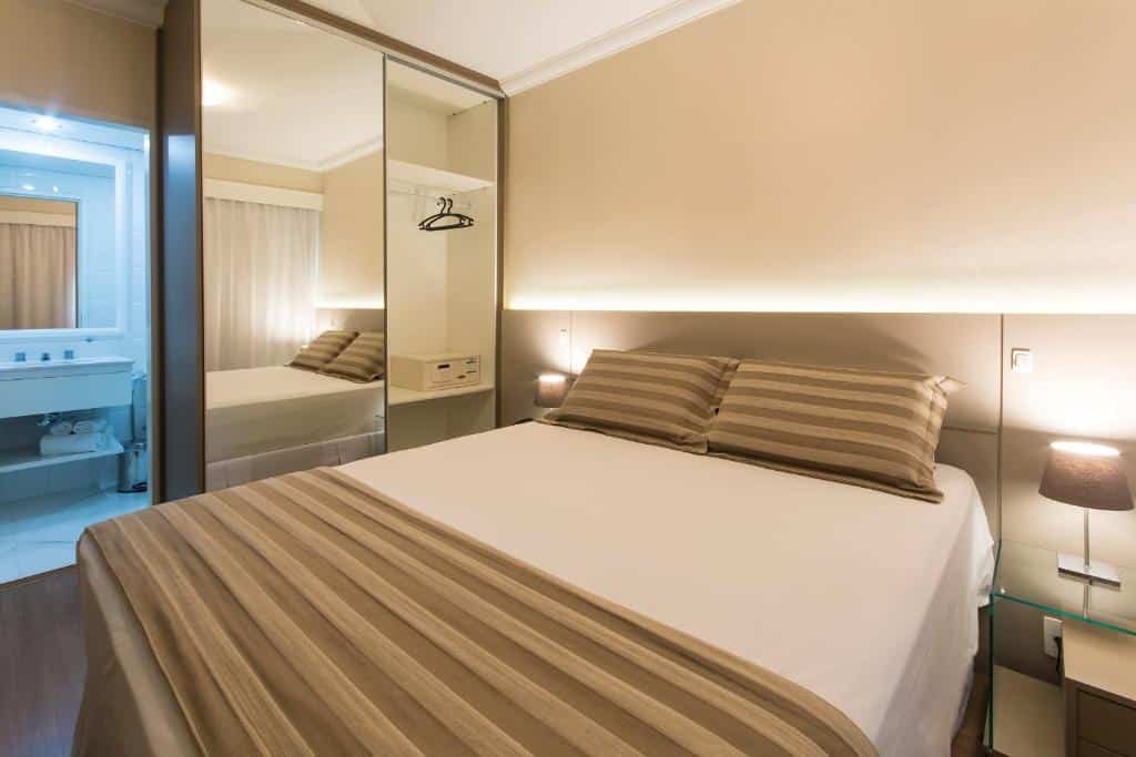 Quarto do Tsue Palace Flat. A cama é box de casal e possui um lençol branco e uma manta bege listrada, os travesseiros também estão da mesma cor e estampa da manta. Há abajures dos dois lados da cama, em uma cabeceira de vidro. Possui um guarda-roupa espelhado encostado na parede. Do lado do móvel, tem a porta que dá acesso ao banheiro. Foto par representar o post sobre hotéis em Moema.