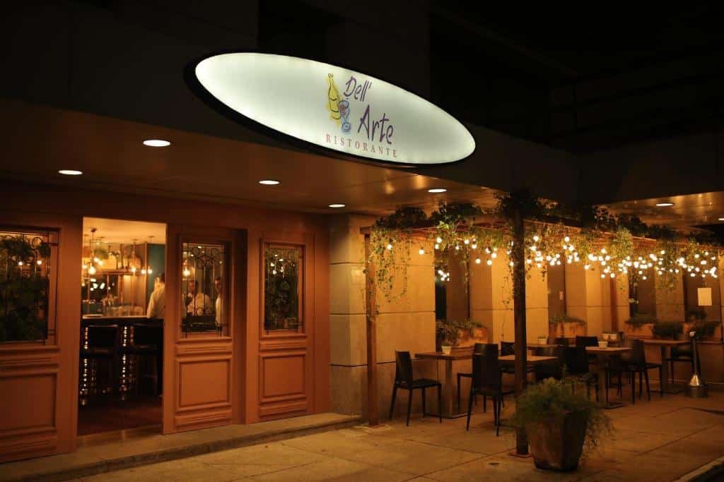 Restaurante italiano no hotel Mercure São Paulo Times Square. Foto tirada à noite. Na área externa do restaurante há várias mesas e cadeiras, e uma iluminação aconchegante com muitas plantas.