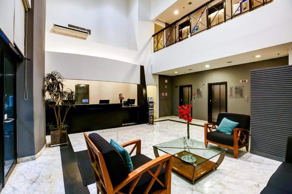 Saguão do hotel Slaviero Ibirapuera. No centro, há duas poltronas pretas suportadas em madeira marrom-alaranjada, e há uma mesa de vidro no centro, com um vaso de vidro com flores vermelhas. Do lado, há o balcão, também preto, da recepção, e um grande vaso de vidro com plantas para decoração à sua frente. Suspenso mais a cima, há um ar condicionado. No lado direito da imagem, vemos o acesso para dois elevadores. Também podemos ter a visão do segundo andar do saguão, protegido por barras pretas. Foto para ilustrar o post sobre hotéis em Moema.