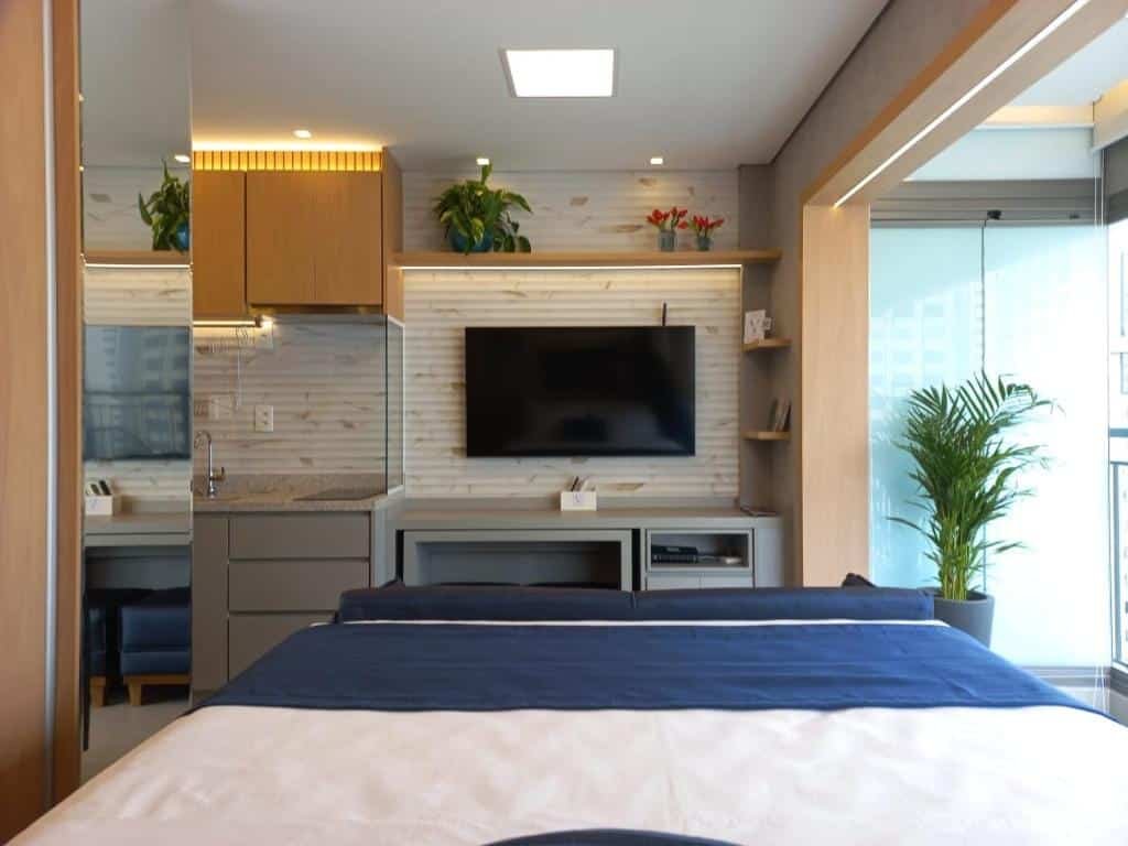 Vista do estúdio em Moema. A foto foi tirada da cama, com visão de frente para a TV, há uma escrivaninha cinza claro debaixo, que comporta gavetas. Ao redor d parede que a TV está fixada, há algumas prateleiras beges. Do lado esquerdo, temos uma visão singela da cozinha. Os armários são em marrom claro alaranjado. Em baixo, podemos ver a pia, na mesma cor da escrivaninha. Do lado esquerdo da cama se encontra o guarda-roupa, na mesma cor dos armários. E, do lado esquerdo da cama, tem uma janela com varanda.