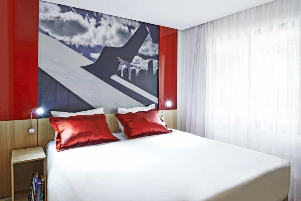 Suíte com janela, cortinas brancas, uma cama de casal com lençol branco, dois travesseiros brancos e dois vermelhos. Há uma pequena cabeceira com alguns livros no lado direito da cama. Em ambos os lados da cama, existem duas luminárias. Na parede, tem um quadro entre duas placas vermelhas que compõem o visual moderno da suíte.