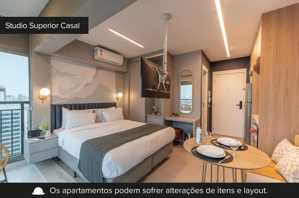 Foto da suíte superior de casal do Charlie Vitrali. A visão que temos do quarto é da cama box de casal, em cores cinzas e com lençol e travesseiros brancos. Há uma mes de cabeceira cinza do lado direito, com um pequeno vaso de planta e o que prece ser um aparelho de internet. Há luminária nos dois lados da cama. No teto, há um ar-condicionado fixado. Do lado esquerdo da cama, há um pequeno armário, um espelho e uma pequena penteadeira com um banquinho. Ainda no quarto, de frente para a cama, podemos ver uma mesa circular com pratos, taças e talheres dispostos. Um sofá amarelo acompanha a mesa. Do lado do sofá, é possível enxergar a cozinha equipada, com gabinetes e armários amadeirados. Na frente da cozinha, há a porta que dá entrada para o banheiro. Por fim, a porta de entrada à suíte. Foto para ilustrar o post sobre hotéis em Moema.