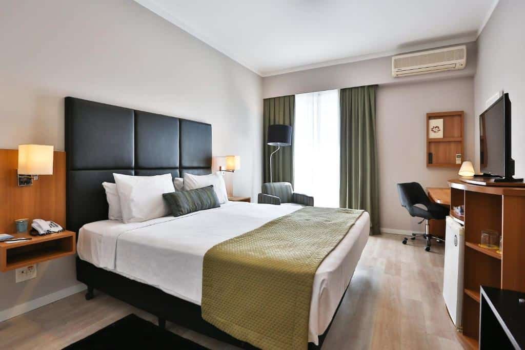 Suíte no Comfort Ibirapuera. O quarto comporta uma cama box de casal, com cabeceira preta, e lençóis brancos. Há alguns travesseiros brancos e um pequeno travesseiro preto no meio. Uma manta bege está posicionada no final da cama. De frente para a cama, há uma pequena estante comportando um TV em cima e um frigobar em baixo. Em ambos lados da cama, há pequenas cabeceiras fixadas na parede, com um abajur em cada, e um interfone. Do lado esquerdo, há uma poltrona cinza com um abajur preto atrás, do lado da janela, com cortinas cinzas. E, do mesmo lado, mais a frente para o canto, há uma escrivaninha de madeira marrom-alaranjada (assim como a estante e as cabeceiras) com uma cadeira preta giratória, e uma prateleira da mesma cor na parede. No alto, na mesma parede da janela, há um ar-condicionado. Foto para representar o post sobre hotéis em Moema.