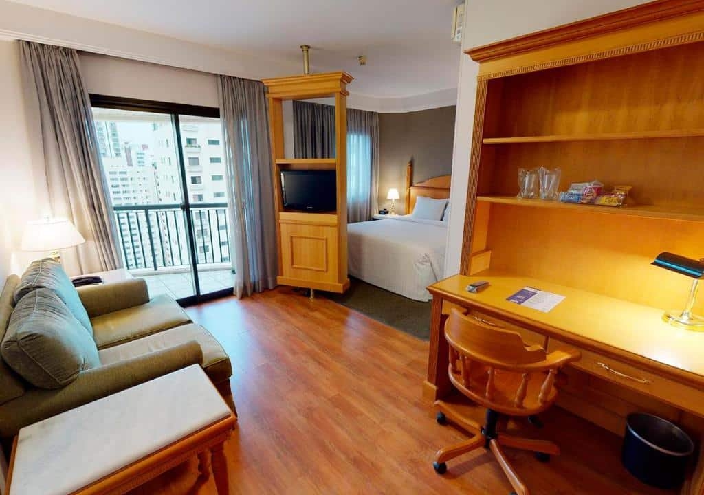 Suíte no hotel Estanplaza. Temos a visão de vários móveis de madeira alaranjada. Possui uma escrivaninha com estantes, gavetas, abajur, cesto de lixo e cadeira (também de madeira). Há um sofá cinza, com um abajur do seu lado esquerdo. O quarto possui varanda. Voltado para o sofá, tem uma televisão suportada em uma estante de madeira, que pode ser girada em diração à cama. Atrás da estante, há uma cama de casal com lençóis brancos. Há uma cabeceira com abajur do lado da cama. Foto para ilustrar o post sobre hotéis em Moema.