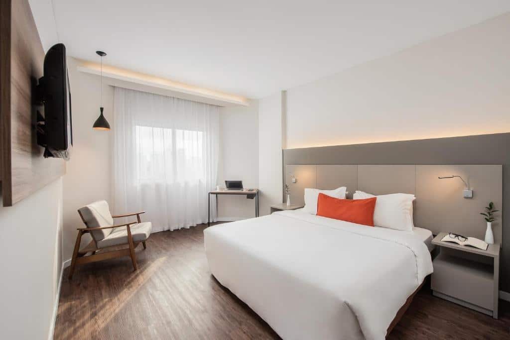 Suíte no Intercity São Paulo Ibirapuera. Cama de csal com lençol branco, travesseiros brancos e um travesseiro vermelho no meio. Há cabeceiras de ambos os lados. De frente para a cama, tem uma televisão fixa na parede. No canto do quarto, há uma poltrona com uma luminária em cima e uma escrivaninha próxima à janela. Foto para ilustrar o post sobre hotéis em Moema.