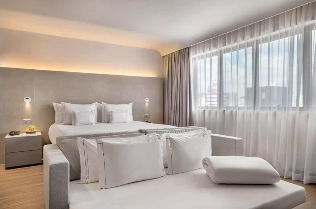 Suíte no Meliá Ibirapuera. As roupas de cama e cortina são brancas. Há um sofá em frente a cama, também na cor branca. A escrivaninha do lado da cama possui o interfone para contato com o staff e um vaso com planta. Os interruptores das luzes estão em ambos os lados da cama. Imagem para representar o post sobre hotéis em Moema.