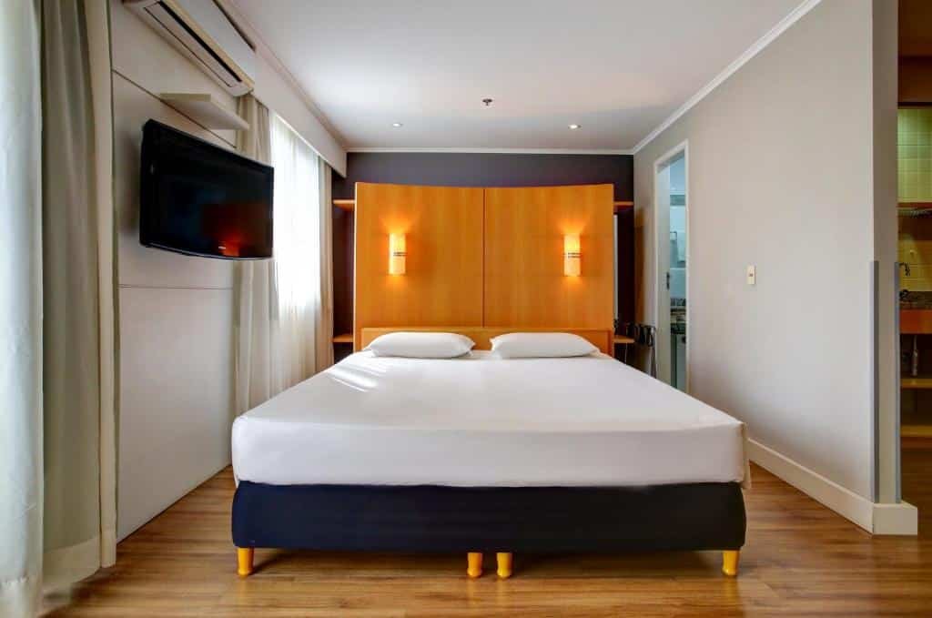 Visão do quarto do hotel Slaviero Ibirapuera. No centro, vemos uma cama de casal com lençóis e travesseiros brancos. A cabeceira é na cor de uma madeira clara de tom alaranjado, suportando duas luminárias de cada lado. Atrás, a parede é azul escura. Do lado direto, tem acesso ao banheiro privativo, do lado esquerdo, uma janela com cortinas claras. Há uma tv suspensa na parede, e acima dela, uma prateleira e um ar condicionado. Foto para ilustrar o post sobre chip internacional para Moema.