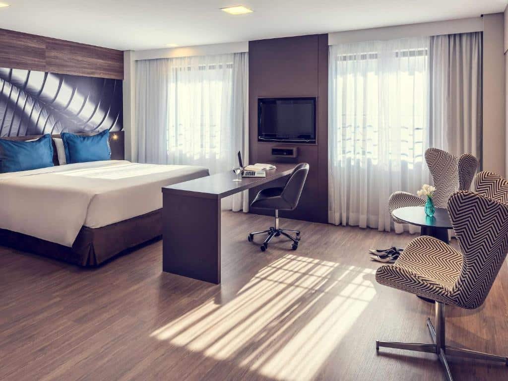 Suíte com cama de casal, lençol branco, travesseiros azuis. As cortinas são brancas, possui duas janelas. Na frente da cama, tem uma mesa com cadeira giratória para facilitar o trabalho com computador. Do lado da mesa, fixo na parede há uma TV. Ainda mais no fundo do quarto há duas poltronas e uma mesa de centro. Quarto bastante espaçoso, imagem utilizada para representar o post sobre hotéis em Moema.