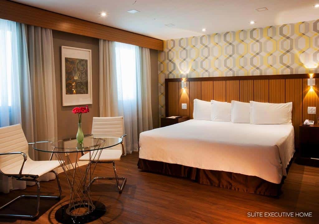 Quarto grande e espaçoso, com uma cama larga para casal. Está bem iluminado, possui janelas e uma mesa de canto com duas cadeiras almofadadas e um vaso com flores em cima. Na escrivaninha ao lado da cama, há um interfone. Foto para representar o post de hotéis em Moema.