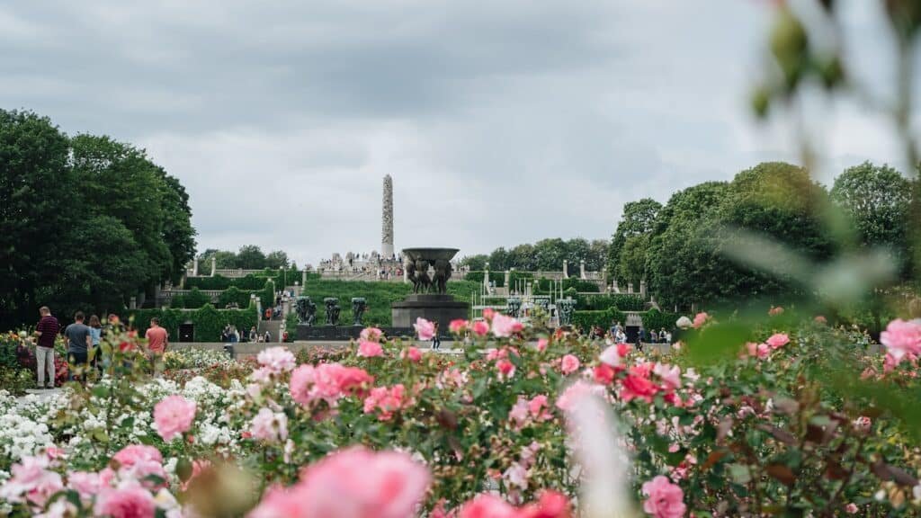 O Vigelandsparken um grande parque com flores rosas desabrochando em primeiro plano, árvores, esculturas e um alto obelisco de pedra ao fundo, que pode ser visitado com o eSIM Oslo.