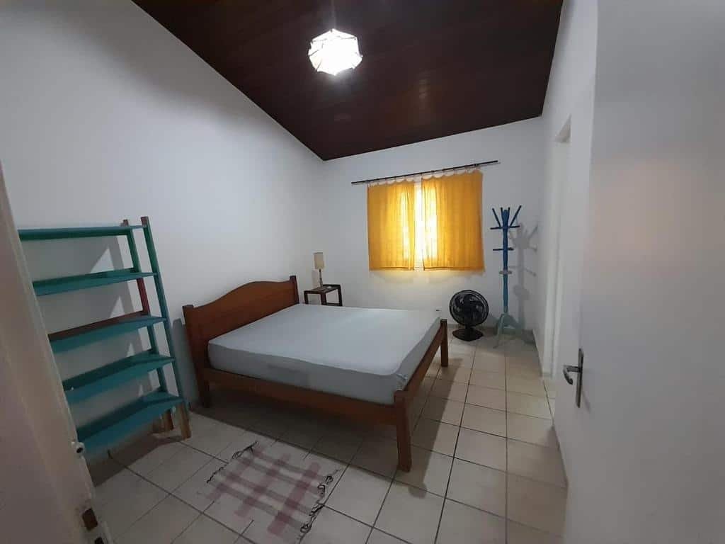Quarto do Aconchego casa. Uma cama de casal está no meio, ao lado de uma grande prateleira. Ao seu outro lado está uma janela, um ventilador e um cabideiro.