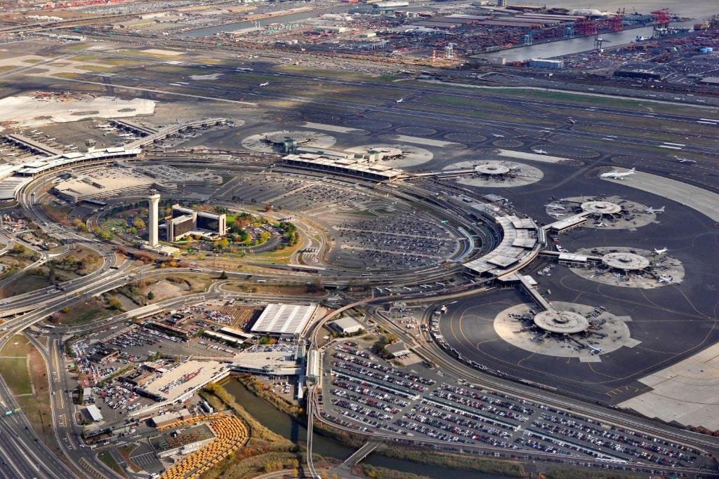 Imagem do Aeroporto Internacional de Newark. A imagem retrata um local amplo e movimentado, com construções imponentes e modernas. Há diversos terminais visíveis, cada um com suas entradas e saídas bem definidas. As pistas de pouso e decolagem se estendem ao longo do aeroporto, com várias aeronaves visíveis em diferentes estágios de preparação ou decolagem. Há também uma área externa com pátios de estacionamento onde as aeronaves aguardam suas partidas.