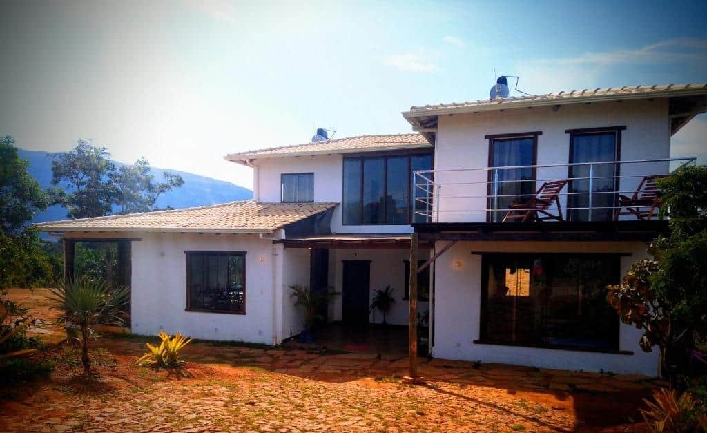 fachada de entrada da pousada aldeia da serra lapinha. É possível ver uma construção de dois andares, pintada de branco, com várias janelas e uma sacada aberta no segundo andar, lado direito da imagem