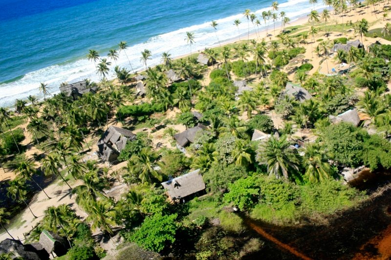 Aldeia Hippie em Arembepe. Várias casinhas simples e caminhos e árvores ao redor, atrás, no fundo, o mar.