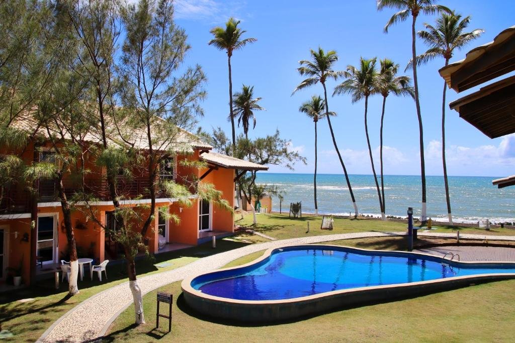 Área externa da Aldeia St. Sebastien 25. Uma piscina no meio, ao redor grama e um caminho de pedras, no lado esquerdo os quartos, no fundo palmeiras e a praia.