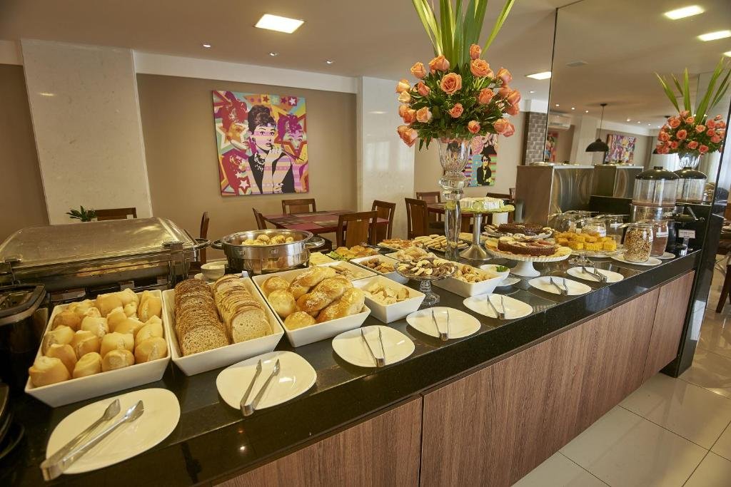 Área de refeições do Alfa Hotel. Há um balcão cheio de comida no meio do saguão e atrás estão mesas com cadeiras.