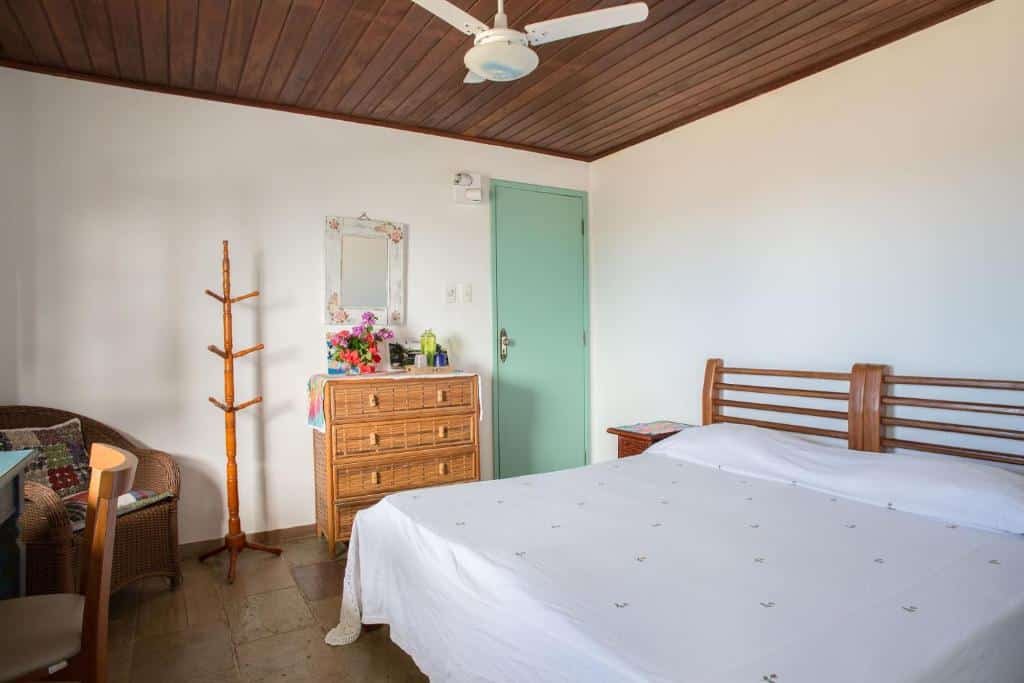 Quarto da Alfazema Cultural Bed and Breakfast. Uma cama de casal no lado direito, no fundo a porta do quarto, uma cômoda, um cabideiro e um espelho. No lado esquerdo uma poltrona e uma mesa de trabalho com cadeira. Bastante luz natural entrando no quarto. Foto para ilustrar post sobre pousadas em Arembepe.