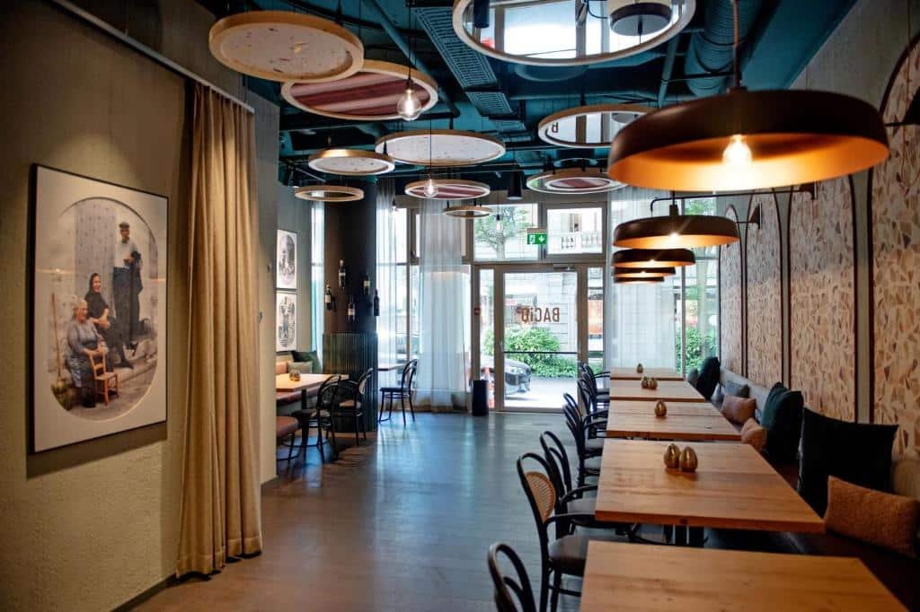 Restaurante no Ameron Luzern. As luzes estão baixas, mas está de dia. Há vários lustres com lâmpadas de cores quentes que passam um ar mais aconchegante. O ambiente possui papéis de parede e mesas quadradas de madeira com sofás e cadeiras. Na parede esquerda, na entrada, há um quadro decorativo. A Porta de entrada está no fundo da foto, é de vidro e é possível ver a rua.