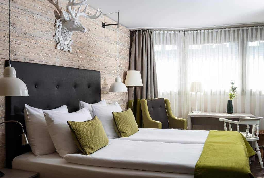 Foto do Ameron Luzern, para representar o post de hotéis em Lucerna. Temos uma visão lateral da cama do quarto, que é box de casal e está encostada na parede esquerda. Possui um abajur que está fixo no teto em seus dois lados. Do lado dela, no fundo do quarto, há uma mesa branca retangular, uma cadeira, poltrona, outro abajur e uma janela na frente.