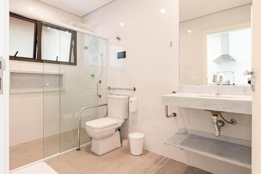 Banheiro acessível do airbnb Anoa Maresias Studios. Do lado esquerdo está o box, com uma barra de abaixo embaixo do chuveiro. Depois dele está o vaso, contornado por duas barras de apoio maiores. Já do lado direito há uma pia com espelho e uma pequena barra de apoio ao seu lado. Representa airbnb na praia de Calhetas
