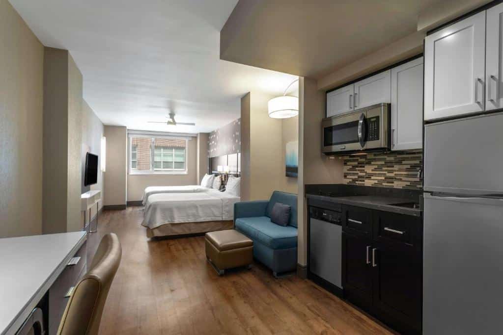 apartamento do Homewood Suites Midtown Manhattan Times Square South com uma cozinha completa no lado direito, onde há uma geladeira, um microondas, uma máquina de lavar louça e um fogão pequeno. No centro do apartamento há um pequeno sofá cama, e mais ao fundo do espaço, há duas camas de solteiro dispostas lado a lado, em frente a televisão