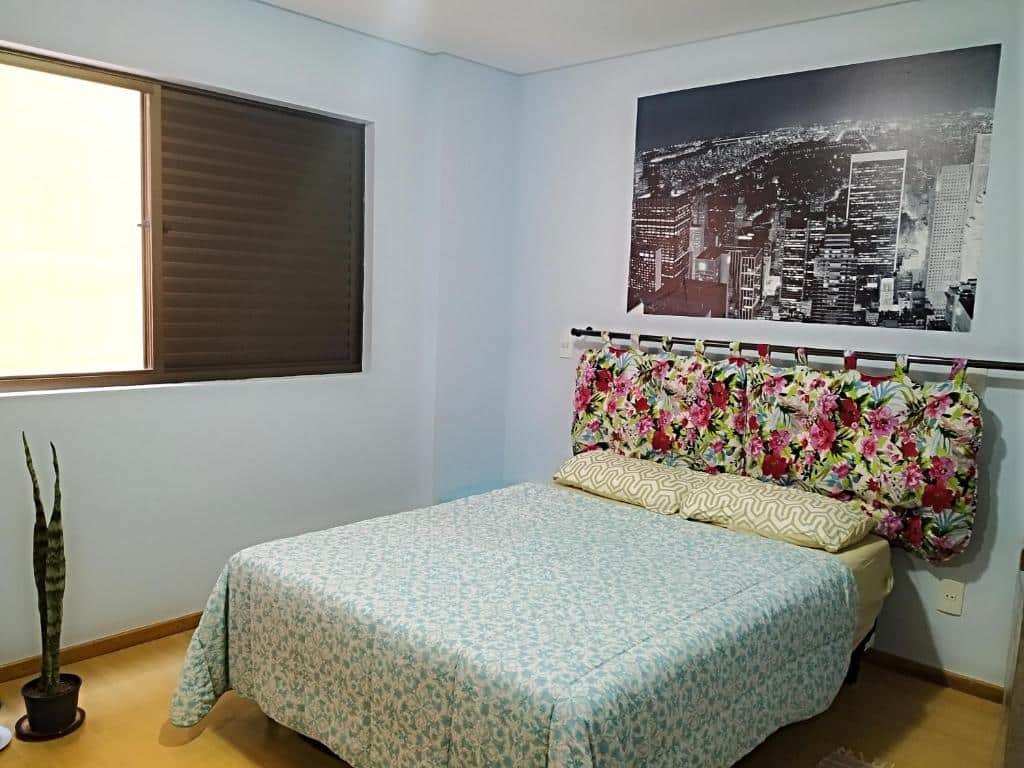 Imagem de um quarto de um apartamento para aluguel de temporada em Viçosa. A cama está centralizada no quarto, há uma cabeceira atrás da cama, uma janela a esquerda e uma pequena planta também a esquerda.