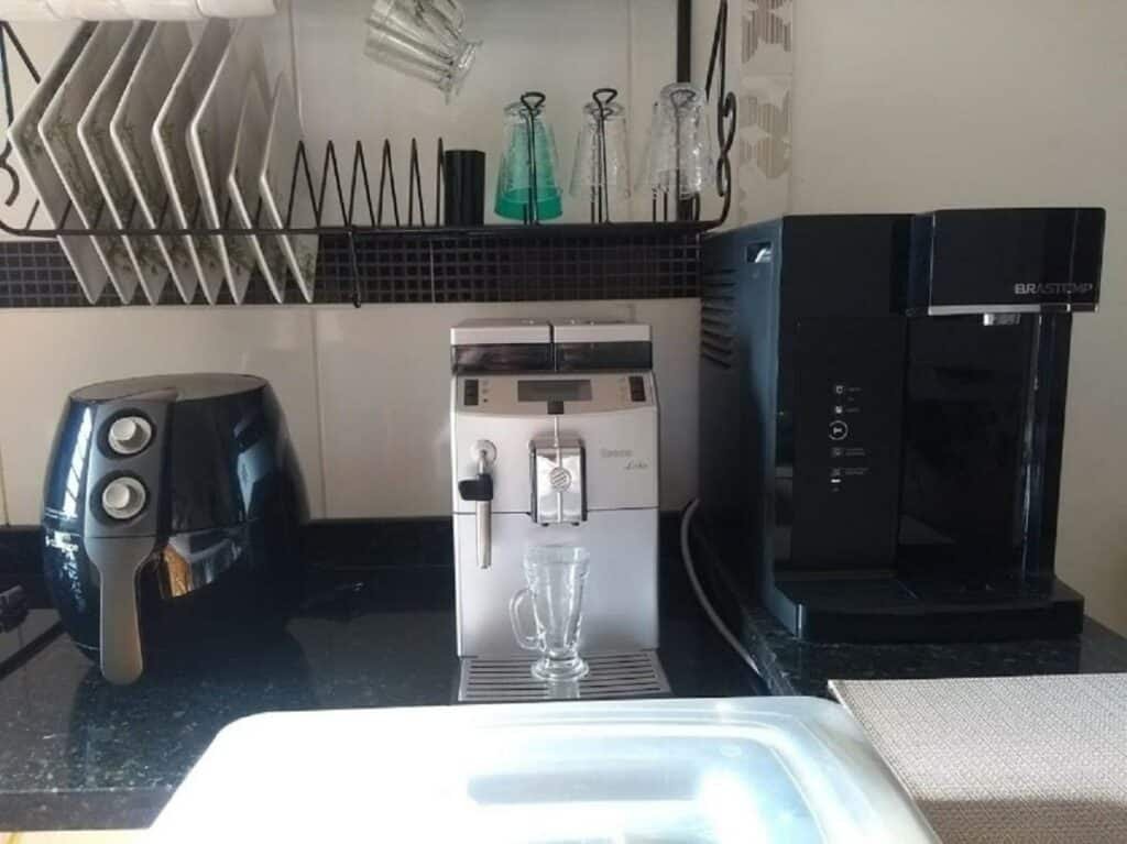 Cozinha do Apartamento Condomínio Boiçucanga. Em um balcão está uma air fryer, um filtro e uma cafeteira. Acima deles estão alguns pratos e copos no escorredor.