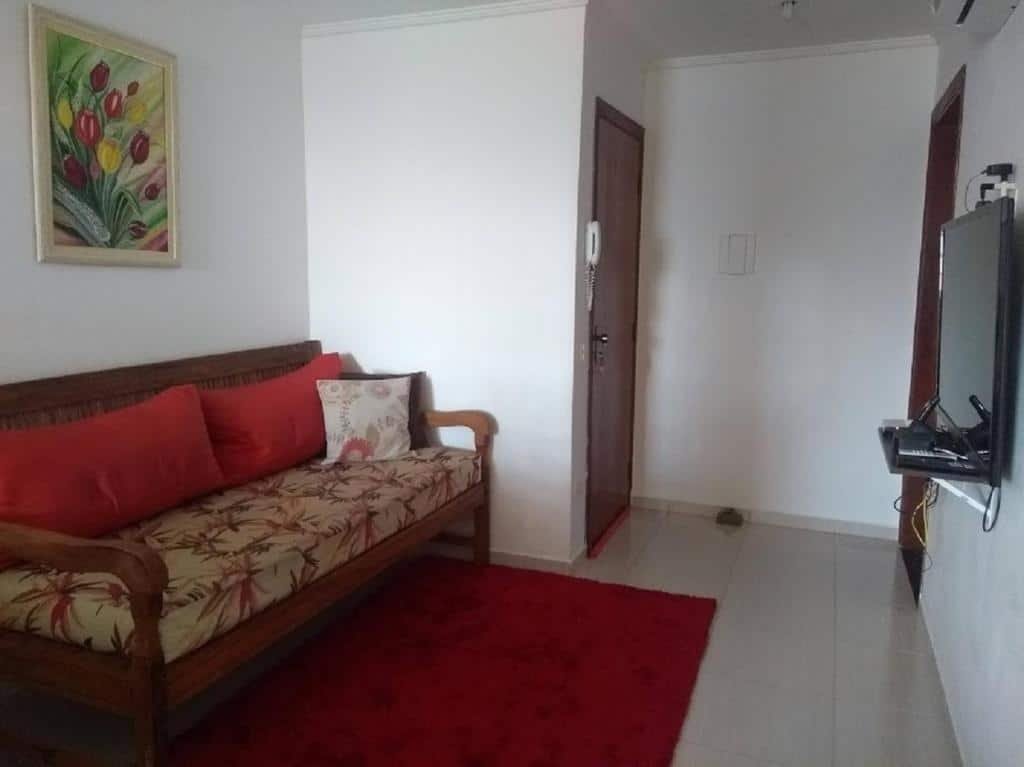 Sala do Apartamento Condomínio Boiçucanga. Um sofá está de frente para uma tv pendurada na parede oposta. No chão entre eles há um tapeta.