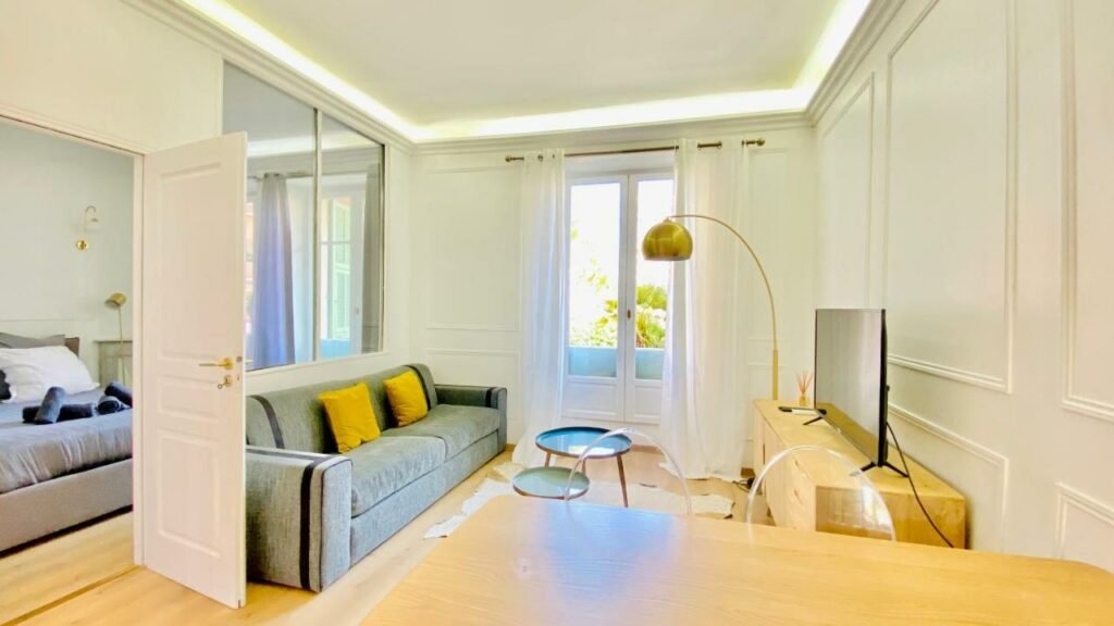 Apartamento Designer Garden View AC, Fiber Wifi Heart of Monaco . Uma mesa na frente, atrás, do lado direito uma televisão, do lado esquerdo um sofá e no centro uma mesinha de apoio. No fundo a porta da sacada. No canto esquerdo a porta do quarto.