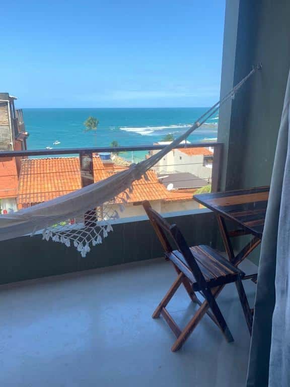 Varanda do Apartamentos de Juan. Do lado direito uma mesa com cadeiras, no meio uma rede de descanso e a vista do mar.