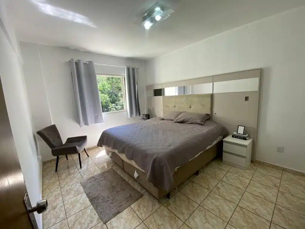 Foto do quarto em Apartamento VIP Centro Serra Negra. O quarto é grande e possui uma cama box de casal com cabeceira espelhada no centro. Na esquerda da cama há uma janela e uma poltrona preta.