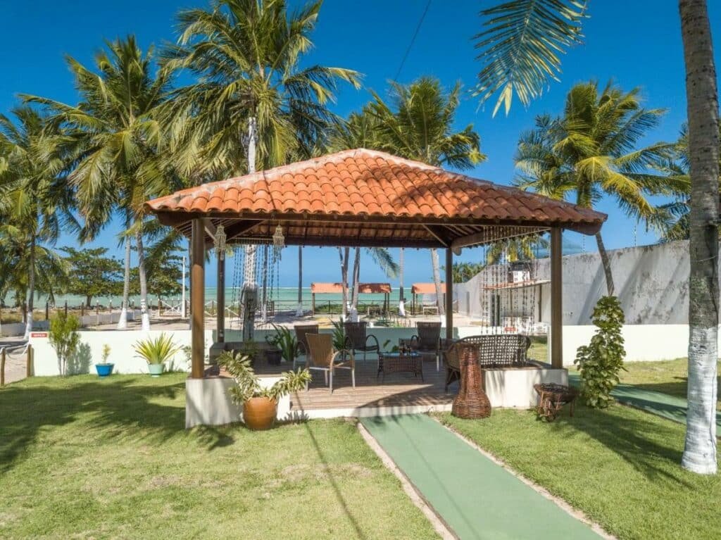 Área externa com um quiosque com cadeiras e um gramado em volta. Há árvores e vasos com flores e o mar ao fundo