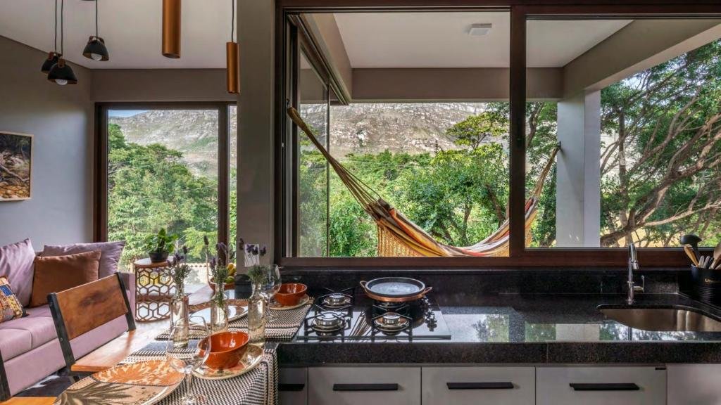 área interna de um dos chalés Aurora da Serra mostrando a cozinha moderna e equipada com janelas amplas que mostram a varanda coberta com rede e vista panorâmica para as montanhas. No canto esquerdo também é possível ver um sofá cor vinho.