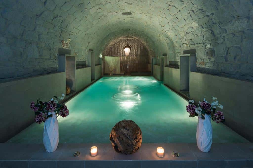 Piscina iluminada, com água cristalina, dentro de um túnel do B2 Boutique Hotel, em Zurique, com teto arredondado e de tijolos cinzas; além de dois vasos de flores roxas e duas velas enfeitando o espaço