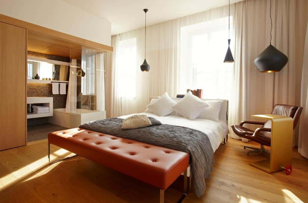 Quarto do B2 Boutique Hotel, com chão de madeira, iluminarias pretas suspensas, poltrona estofada de couro marrom, com uma mesa de madeira ao lado, e banheiro com parede de vidro e hidro dentro