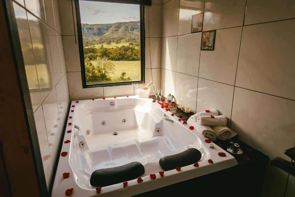 A imagem mostra uma banheira de hidromassagem localizada no Chalé Sol Nascente, decorada com pétalas de rosas ao redor da borda. A banheira está em um espaço azulejado, com uma janela grande que oferece uma vista para uma paisagem natural com montanhas e vegetação. Ao lado da banheira, há toalhas brancas dobradas e itens decorativos, como taças de champanhe e velas, criando um ambiente romântico e relaxante. A iluminação natural que entra pela janela realça a sensação de tranquilidade e conforto.