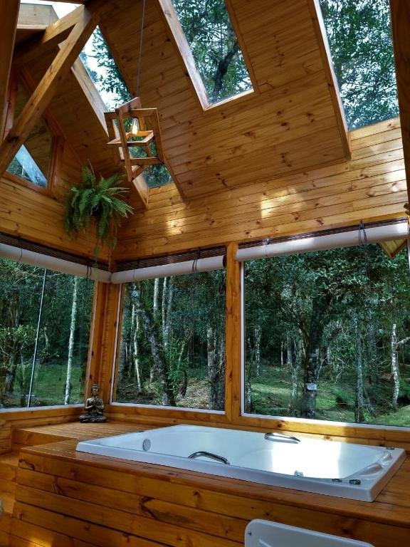 A imagem mostra uma banheira de hidromassagem no Chalés Estância Cachoeira do Avencal, dentro de uma cabana, rodeada por amplas janelas de vidro que oferecem uma vista panorâmica para uma floresta densa e verdejante. As paredes e o teto da cabana são revestidos em madeira de tom claro, criando uma atmosfera aconchegante e rústica. O teto possui claraboias que permitem a entrada de luz natural, destacando a beleza do ambiente. Pendurada no teto, uma pequena luminária de estilo rústico adiciona charme ao local. Uma planta suspensa e uma pequena estátua decorativa ao lado da banheira completam o espaço, proporcionando um ambiente relaxante e intimista em plena conexão com a natureza.