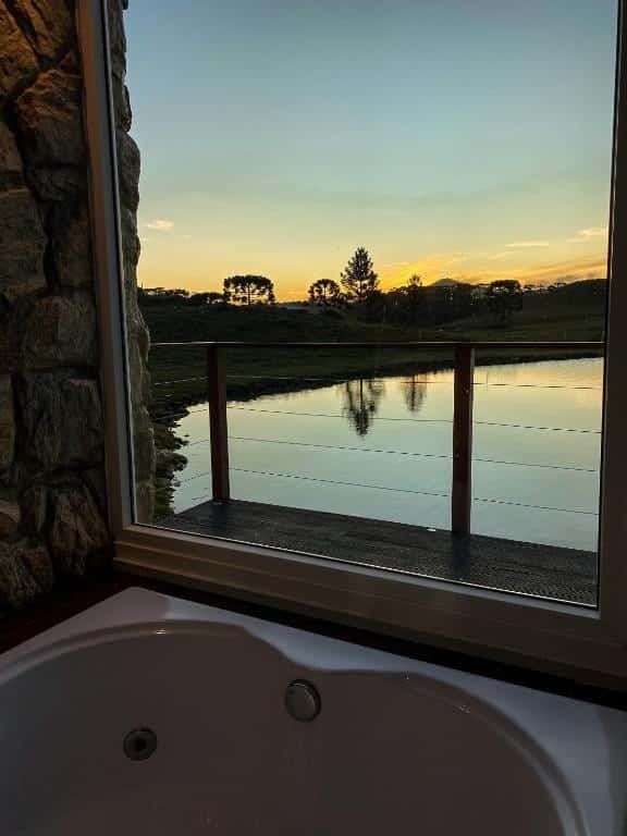 A imagem mostra uma banheira no Recanto Entre Rios posicionada perto de uma grande janela de vidro, com uma vista panorâmica para um lago tranquilo ao entardecer. A parede de pedra ao lado da janela complementa o ambiente rústico e aconchegante. Do lado de fora, o deck de madeira cercado por uma grade com cabos de aço oferece uma vista deslumbrante do lago e da paisagem serrana, com árvores espalhadas e o céu pintado em tons suaves de laranja e amarelo à medida que o sol se põe.