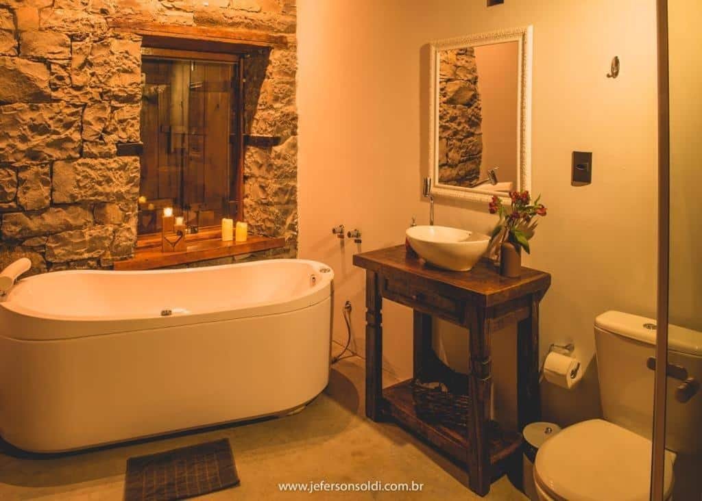 banheiro da Casa Vêneto, com um vaso sanitário, pia, banheira individual, parede de pedra e janela com apoio para velas