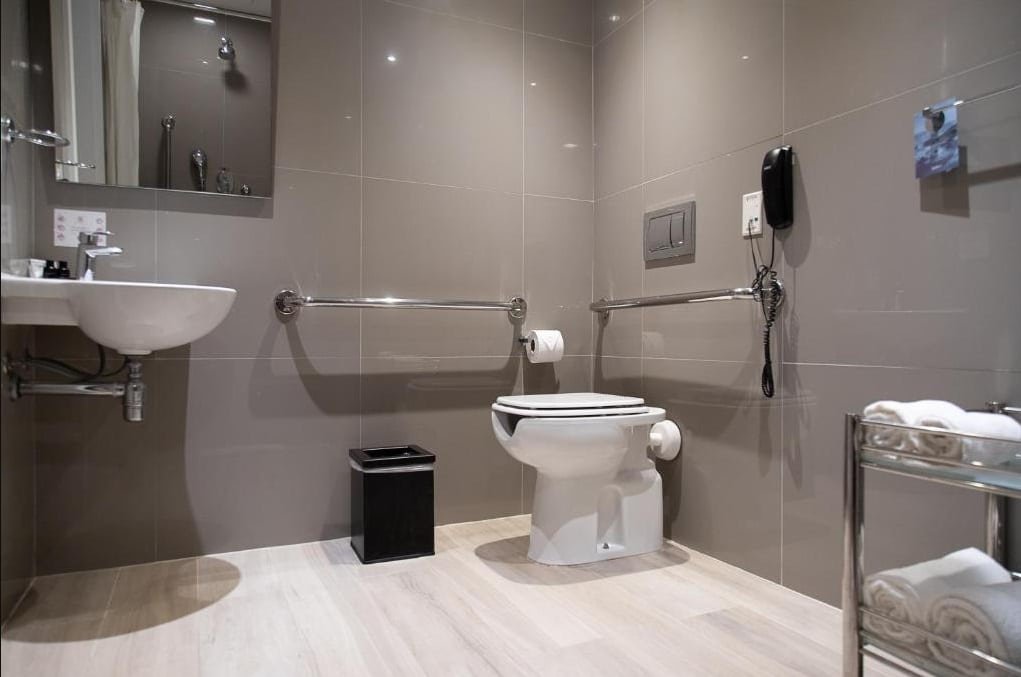 Banheiro adaptado do Radisson Barra com barras de apoio, vaso elevador e pia mais baixa, com espelho e um móvel com toalhas brancas