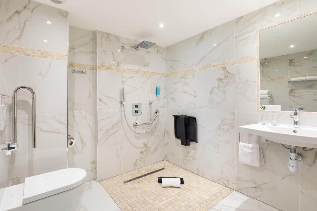 Banheiro do Best Western Hotel Lakmi Nice. No lado esquerdo há uma privada com uma barra de apoio na parede. Já no centro há um chuveiro sem box de vidro com barras de apoio na parede e uma cadeira para melhor acessibilidade. No lado direito há uma pia com espelho em cima e uma toalha de mão mais baixa para melhor acessibilidade também. Imagem para ilustrar o post hotéis em Nice.